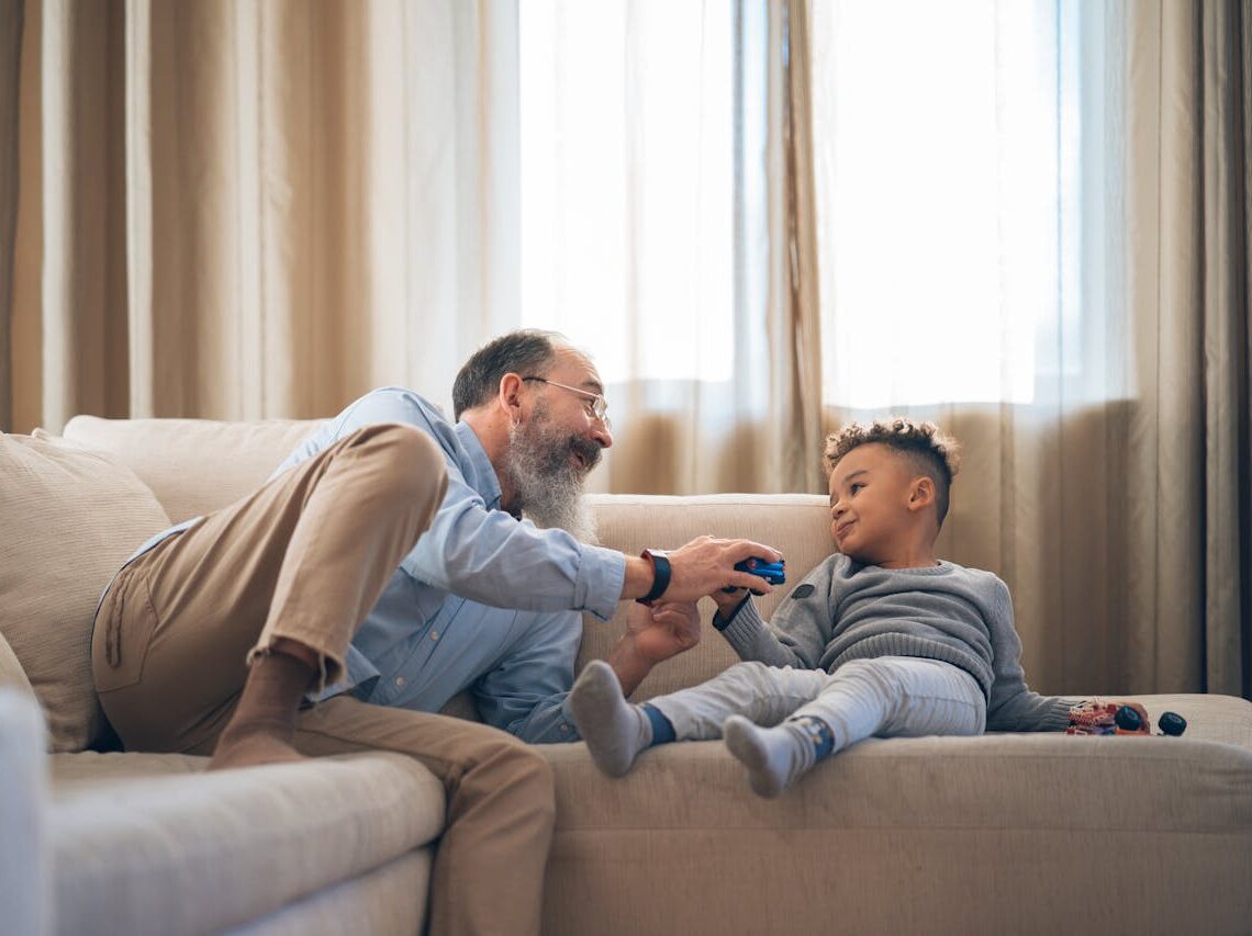 A heartwarming moment between grandfather and grandson laughing and playing on a cozy sofa indoors.